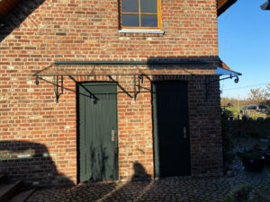 Ein handgeschmiedetes Glasvordach mit grüner Beschichtung, freitragend montiert an einer Backsteinwand. Außerdem zu sehen: ein braunes Fenster im Giebel des Daches sowie zwei grüne Türen.
