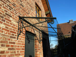 Ein handgeschmiedetes Glasvordach mit grüner Beschichtung, freitragend montiert an einer Backsteinwand. Außerdem zu sehen: ein braunes Fenster im Giebel des Daches sowie zwei grüne Türen. Im Hintergrund ein weiteres Gebäude und der Blaue Himmel.