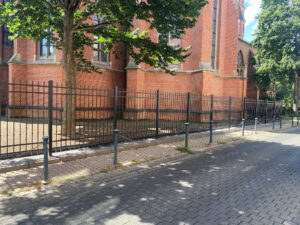 Moderner, vernieteter Metallzaun mit kunstvollen Zierelementen, aufgestellt vor einer Backsteinkirche.