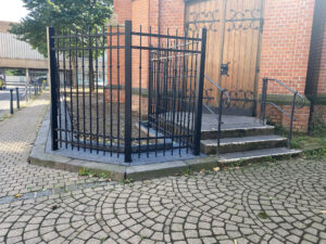 Detailansicht eines hochwertigen Kunstschmiedezauns mit Zierelementen und Nieten vor dem Backsteinmauerwerk einer Kirche.