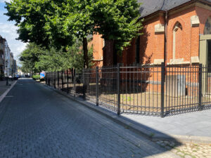 Foto eines kunstvoll gearbeiteten Metallzauns mit Nieten und Zierelementen, montiert vor einer roten Backsteinkirche.