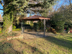 Zu sehen ist ein handgefertigtes Kupferdach im Zentrum des Fotos, umgeben von grünen Bäumen und Hecken. Durch die Zweige scheint der blaue Himmel und betont die filigranen Details dieser kunstvollen Überdachung. Dieses Projekt zeigt, wie individuelle Vordächer und Kunstschmiedearbeiten stilvoll mit der Natur harmonieren und langlebige Akzente am Gebäude setzen.