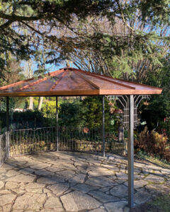 Aus einem anderen Blickwinkel ist hier das kunstvoll gearbeitete Kupferdach dieser Überdachung zu sehen, das elegant von grünen Hecken und hohen Bäumen eingerahmt wird. Die filigranen Schmiedearbeiten kommen besonders gut im Zusammenspiel mit den natürlichen Farben zur Geltung.