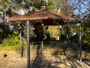 Diese Perspektive zeigt die handgeschmiedete Kupfer-Überdachung von der Seite, eingebettet in üppiges Grün. Die kunstvollen Details und das warme Metall harmonieren perfekt mit der Backsteinfassade und schaffen einen einzigartigen Blickfang für Eingangsbereiche.